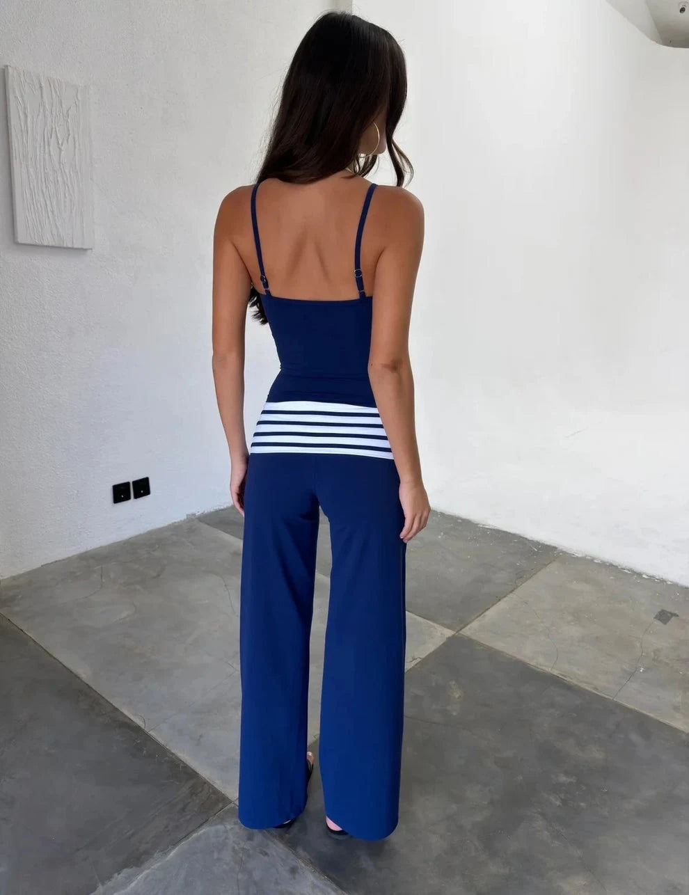 A woman wearing a navy blue sleeveless jumpsuit with thin straps, a fitted bodice, and wide legs. The jumpsuit has a distinctive horizontal band of white stripes around the waist.