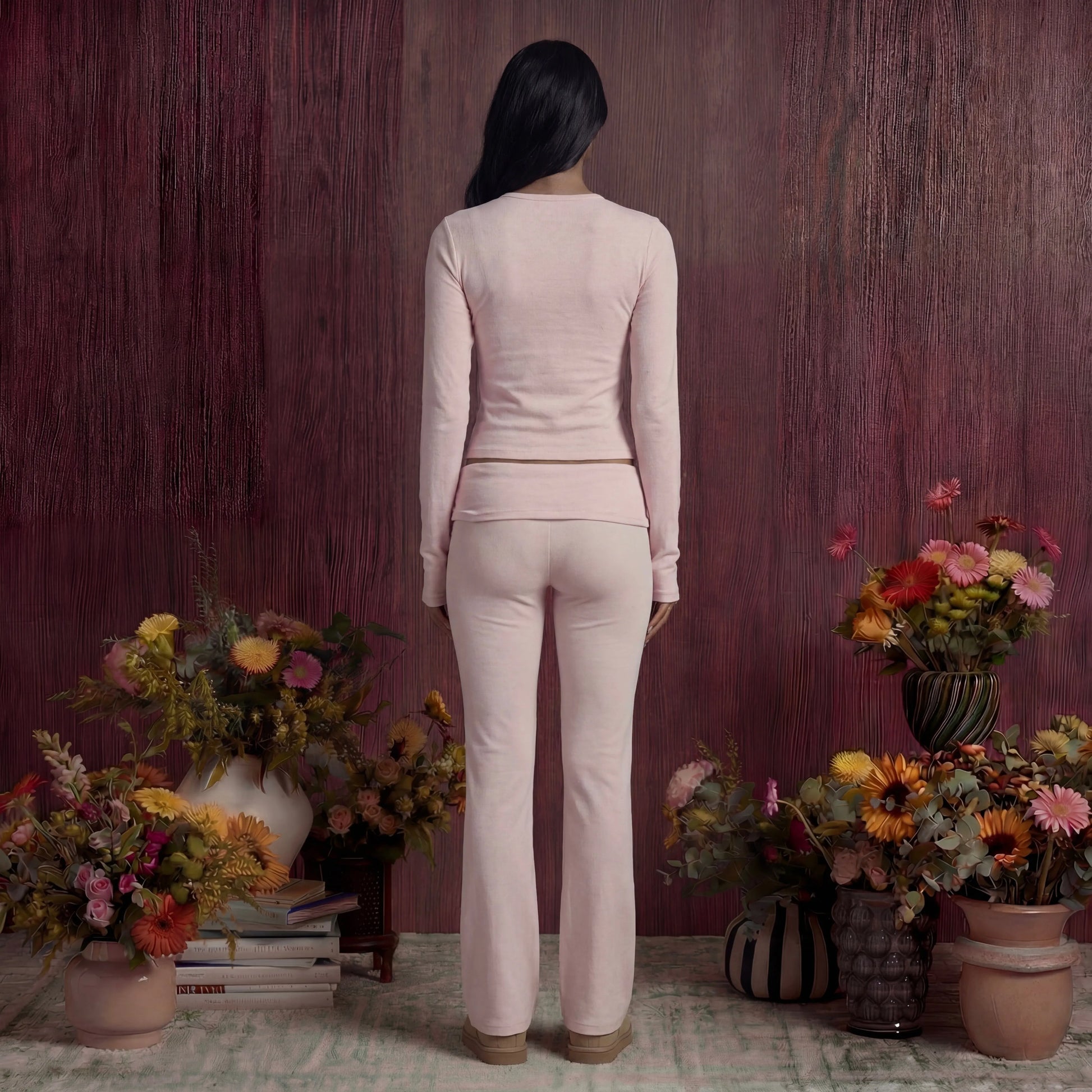 Woman in a light pink cotton fold over pants and top standing in front of a wooden wall with floral arrangements.