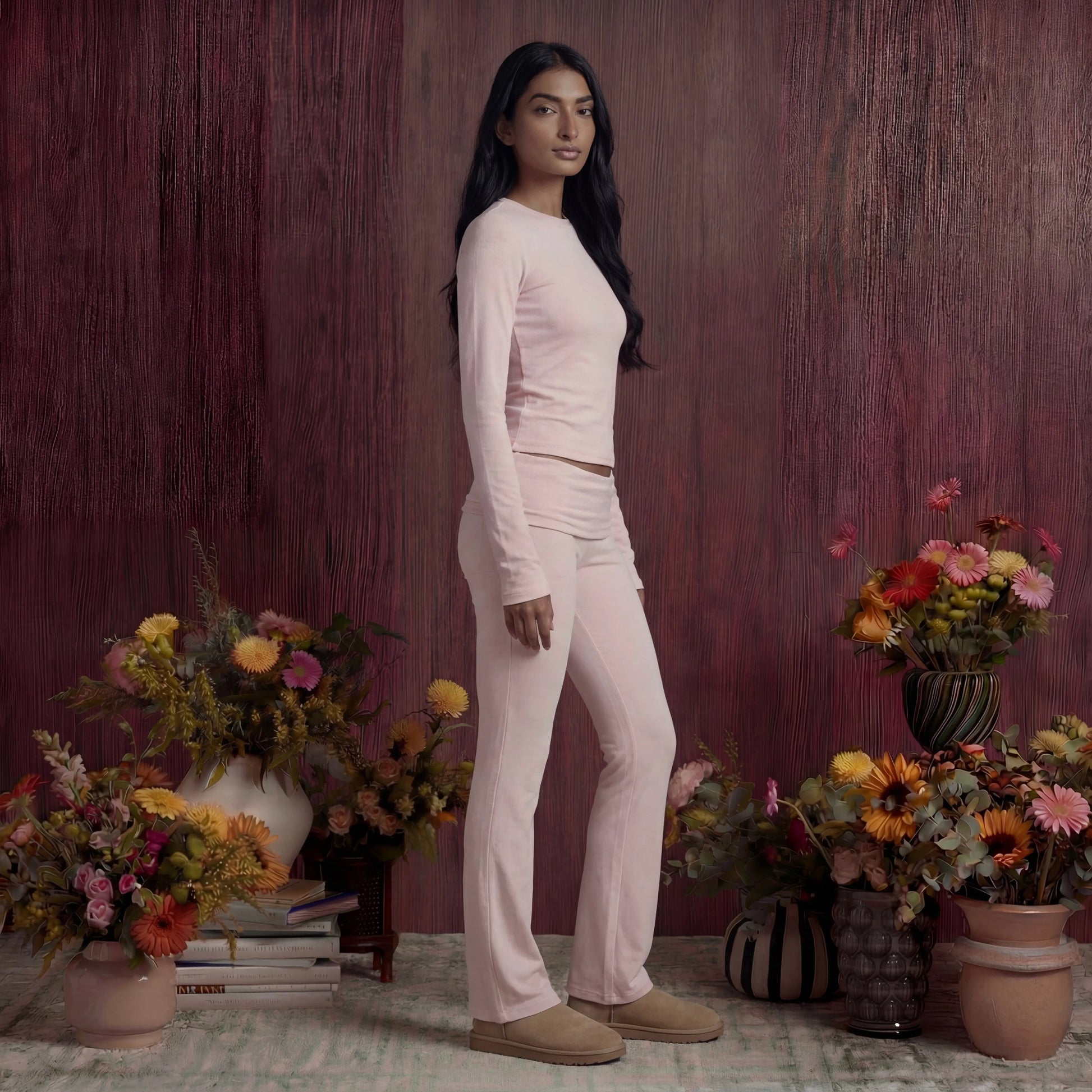 Woman in a white cotton fold over pants and top standing among potted flowers against a textured purple wall.