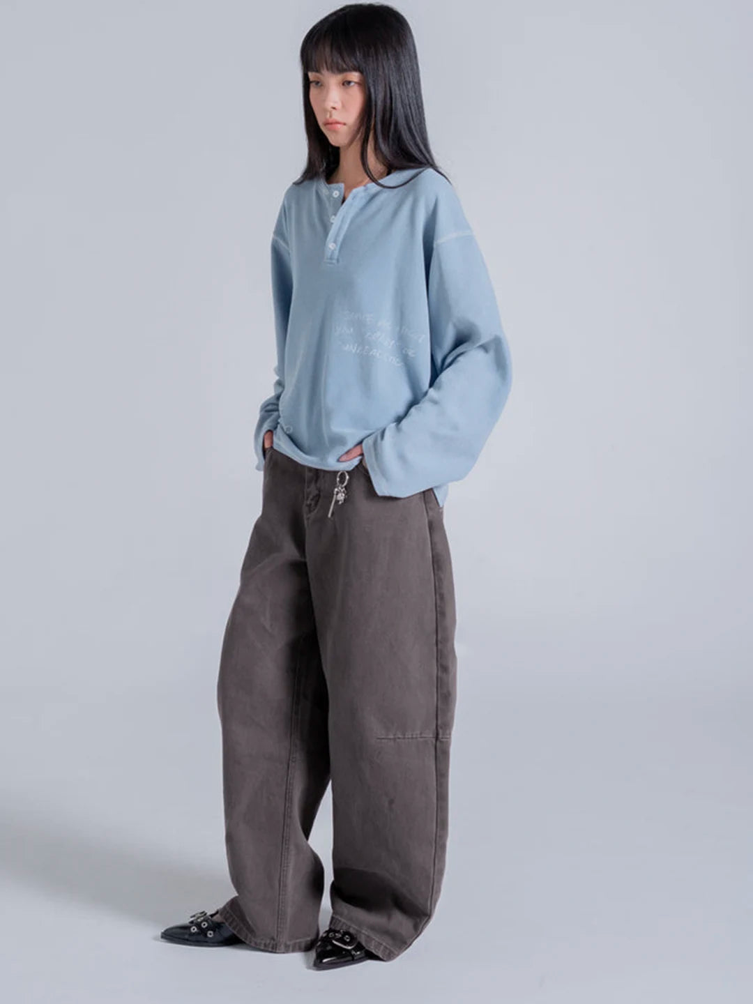 A model wearing a light blue long-sleeve henley-style t-shirt with subtle white text on the chest, paired with wide-leg brown pants and black pointed shoes.