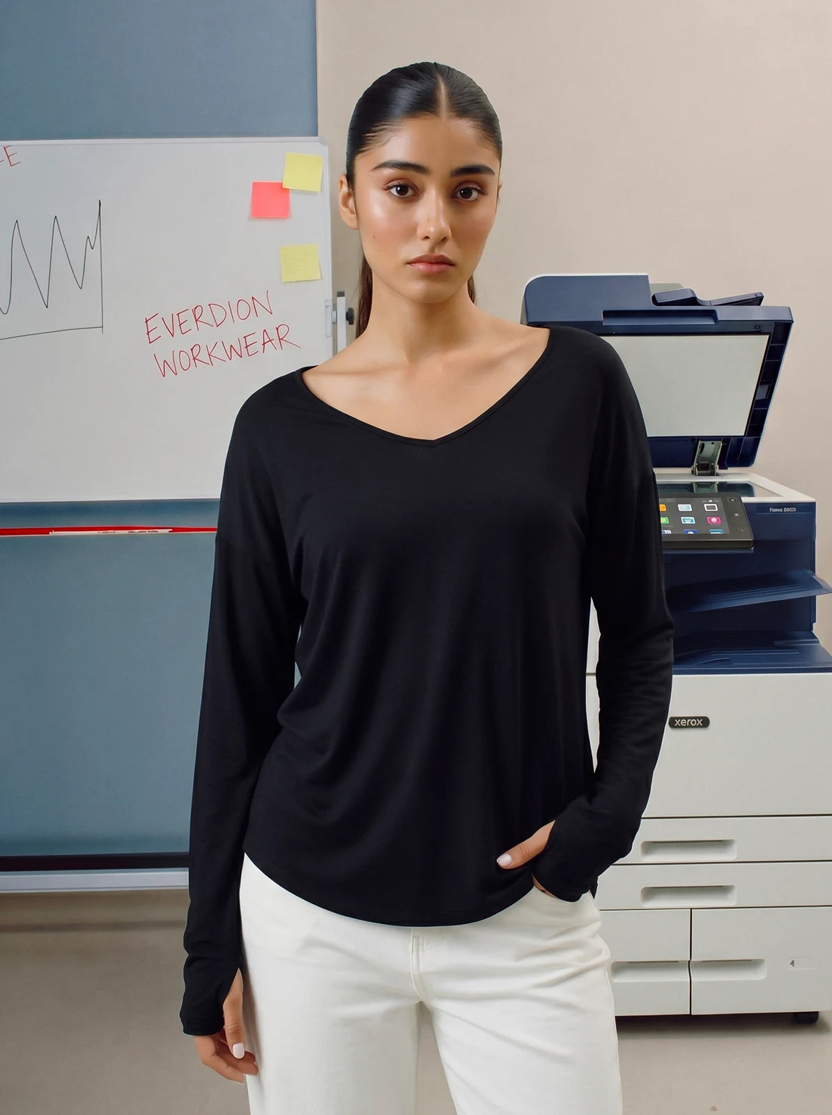Woman wearing a black long-sleeve top and white pants standing in an office setting.