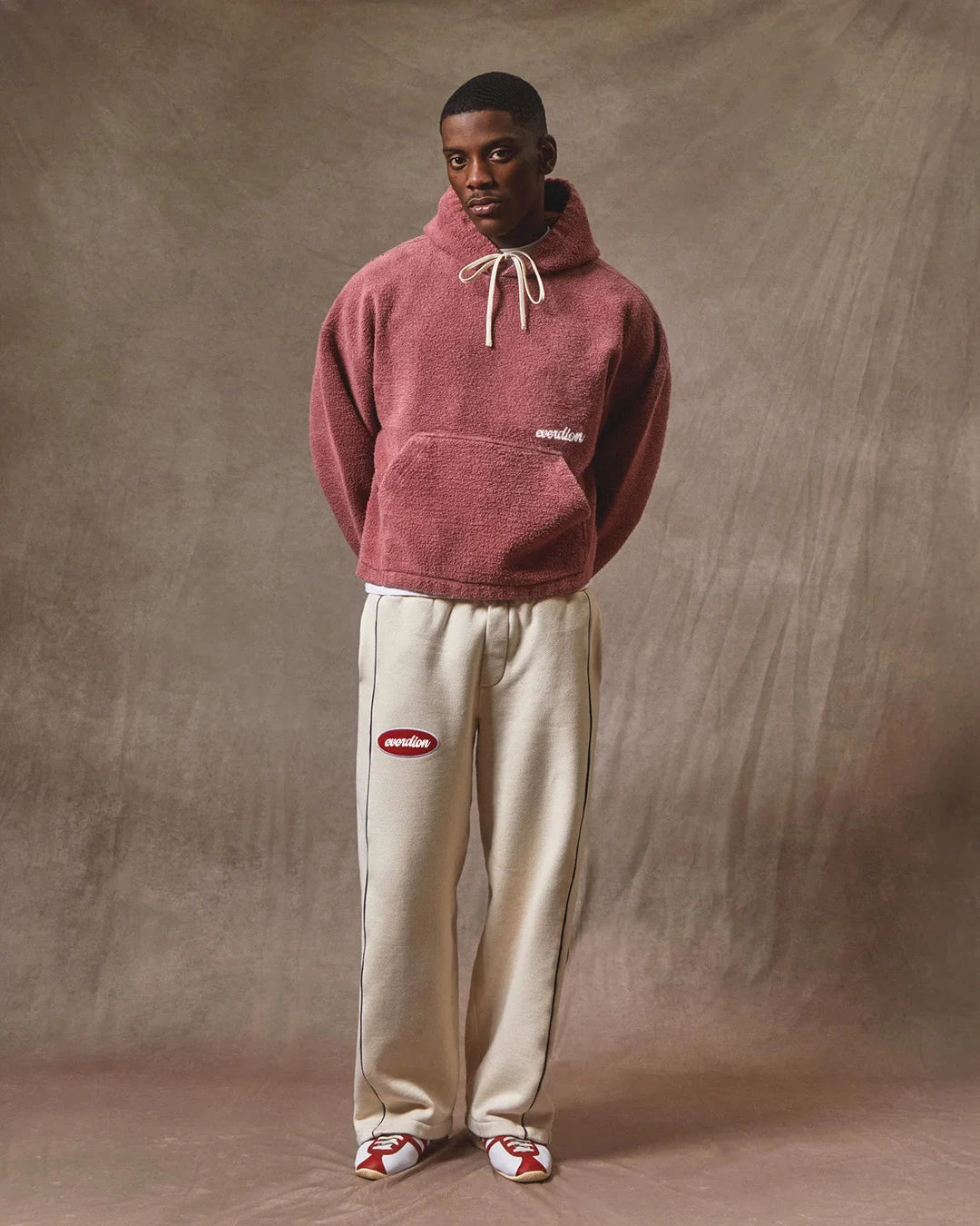 Person wearing a maroon hoodie and beige pants with a brand logo against a brown background