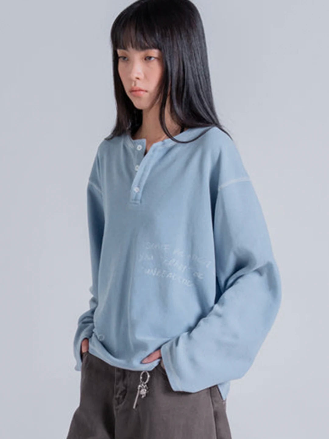 A model wearing a light blue long-sleeve henley t-shirt with white embroidered text on the front, paired with dark brown pants