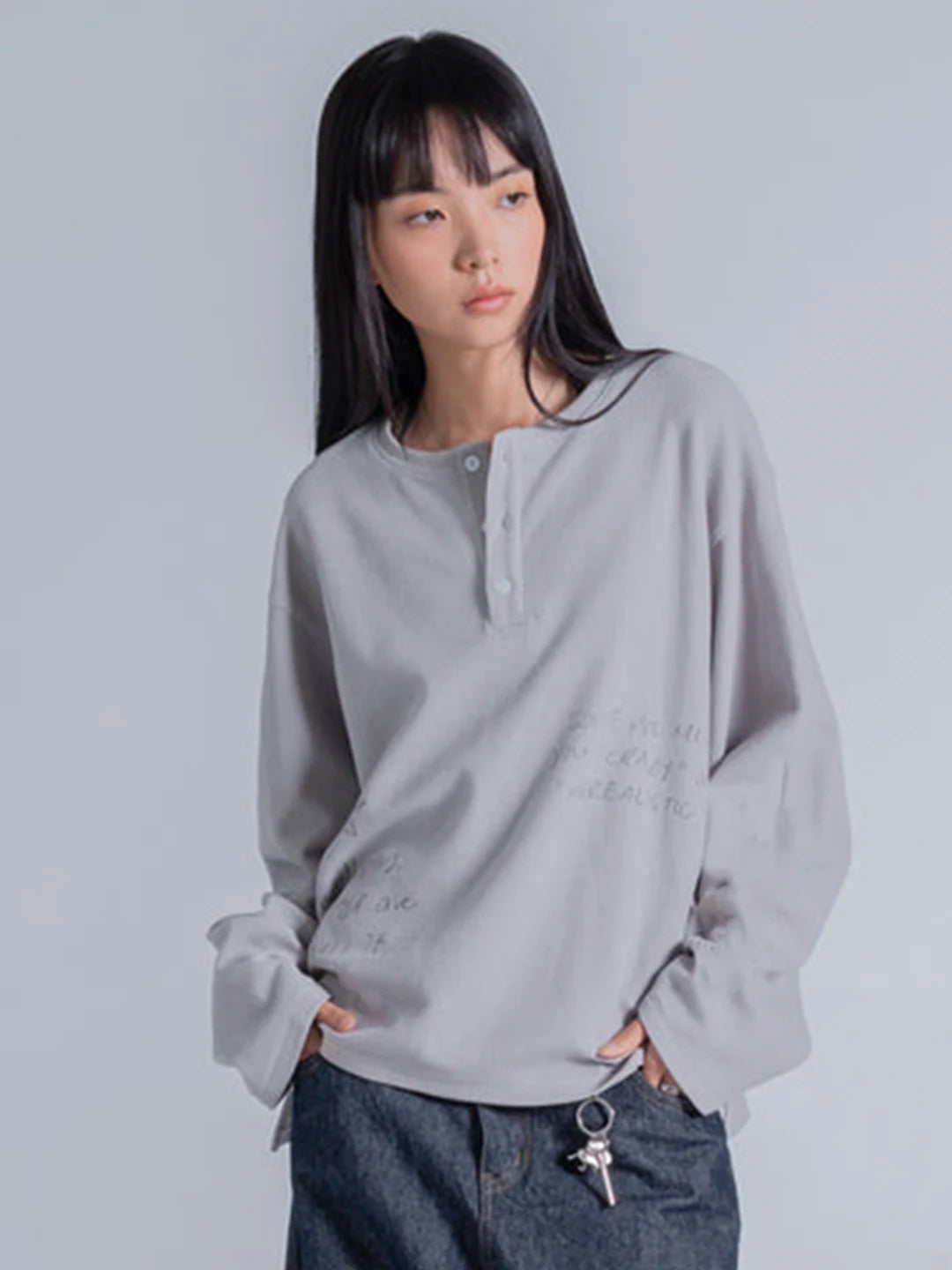 A model wearing a light grey long-sleeve henley shirt with subtle script text on the front, paired with dark blue jeans