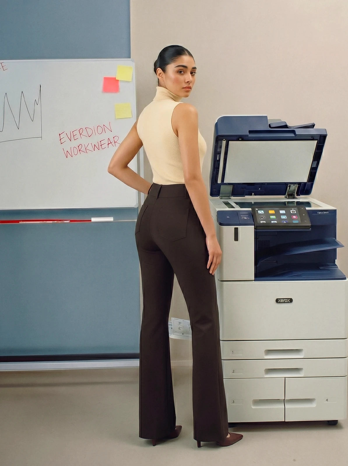 Person wearing brown pants standing in an office setting with a printer and whiteboard in the background.