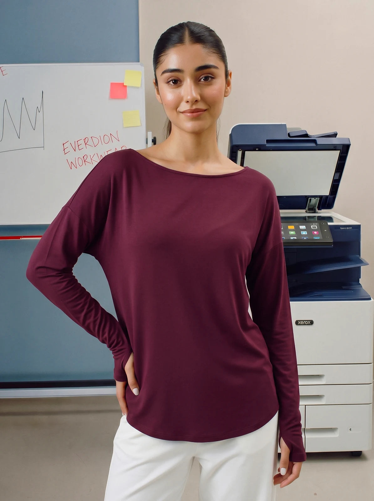Woman wearing a maroon long-sleeve top standing in an office setting with a printer and whiteboard in the background.