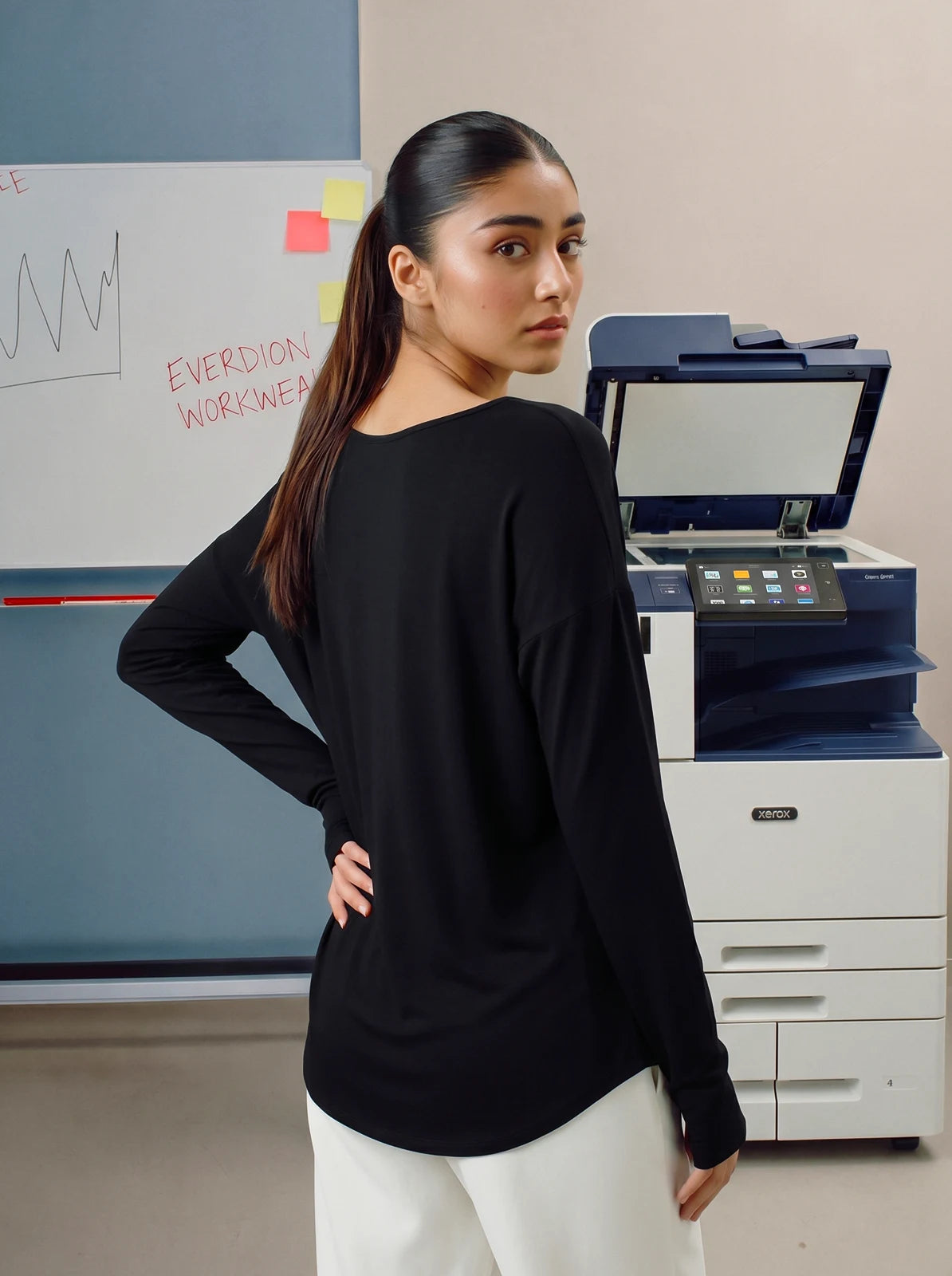 Woman wearing a black long-sleeve top and white pants standing in an office setting.
