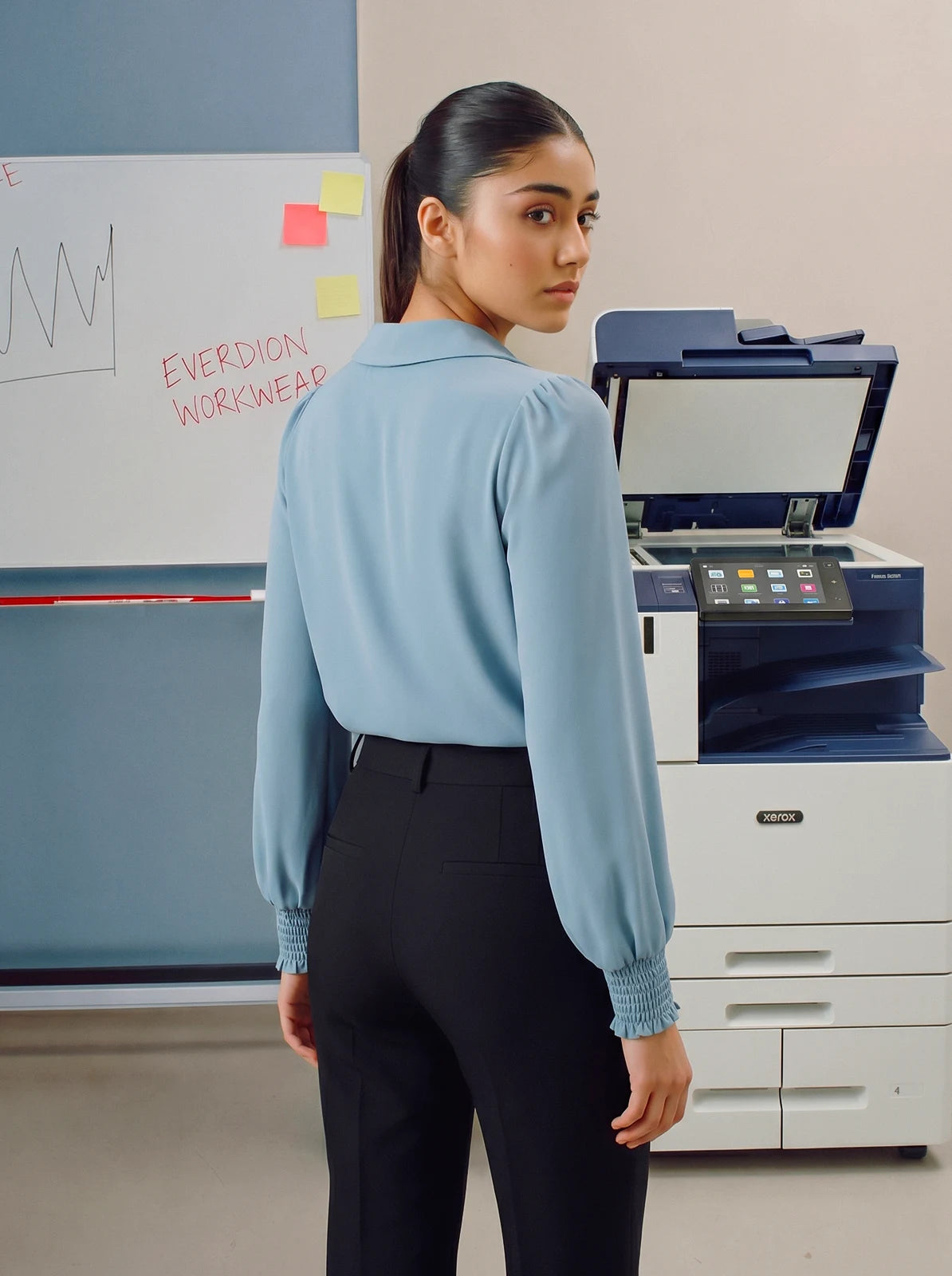 Person wearing a light blue blouse standing in an office setting with a printer and whiteboard in the background.