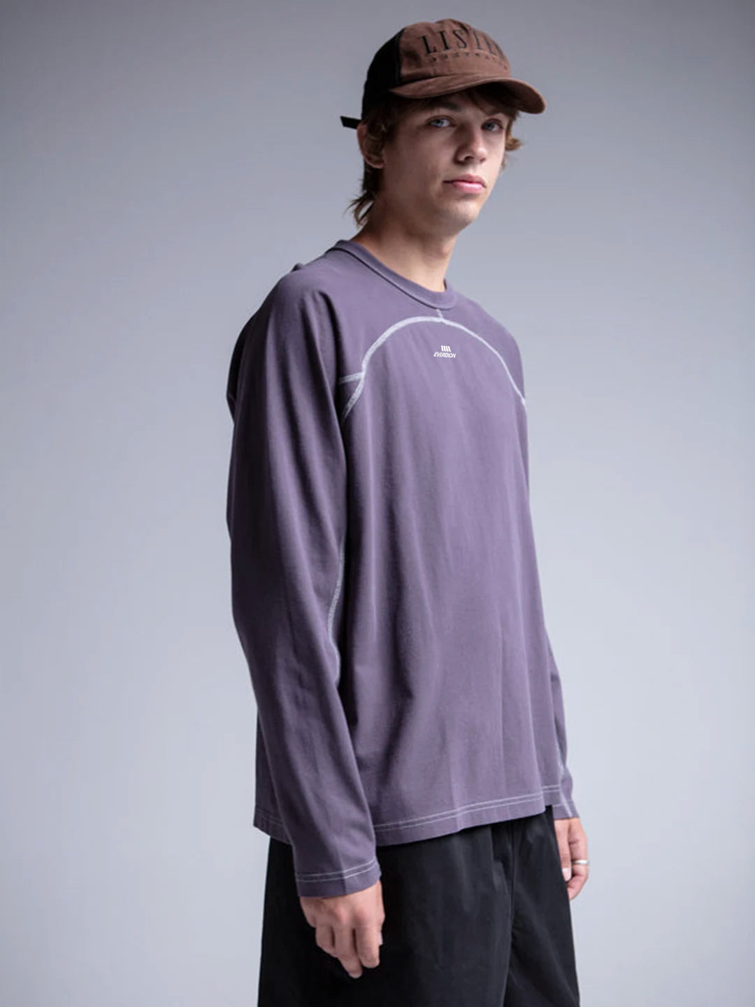 A model wearing a lavender long-sleeve crewneck t-shirt with white contrast stitching and a small white chest logo, paired with black pants and a brown cap