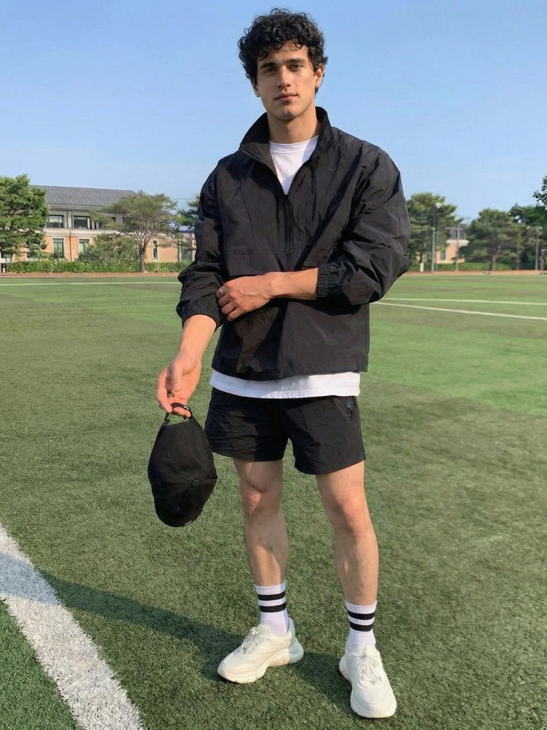 Person standing on a grassy field holding a cap, wearing a black jacket and shorts.