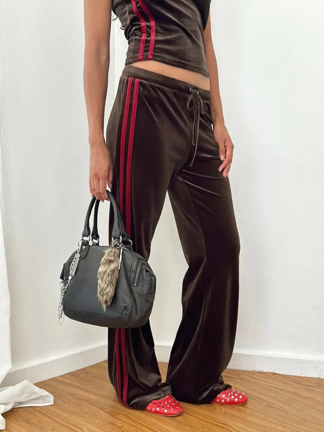 Person wearing brown tracksuit with red stripes holding a gray handbag indoors.
