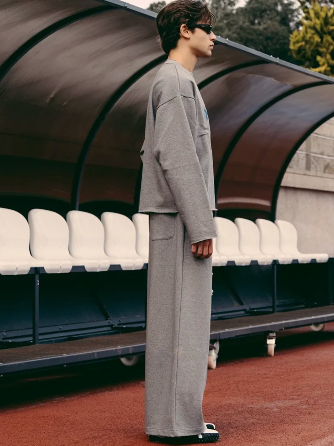 A man wearing a matching grey knit lounge set consisting of a cropped long-sleeve top and wide-leg trousers, standing in profile in front of an outdoor stadium seating area.