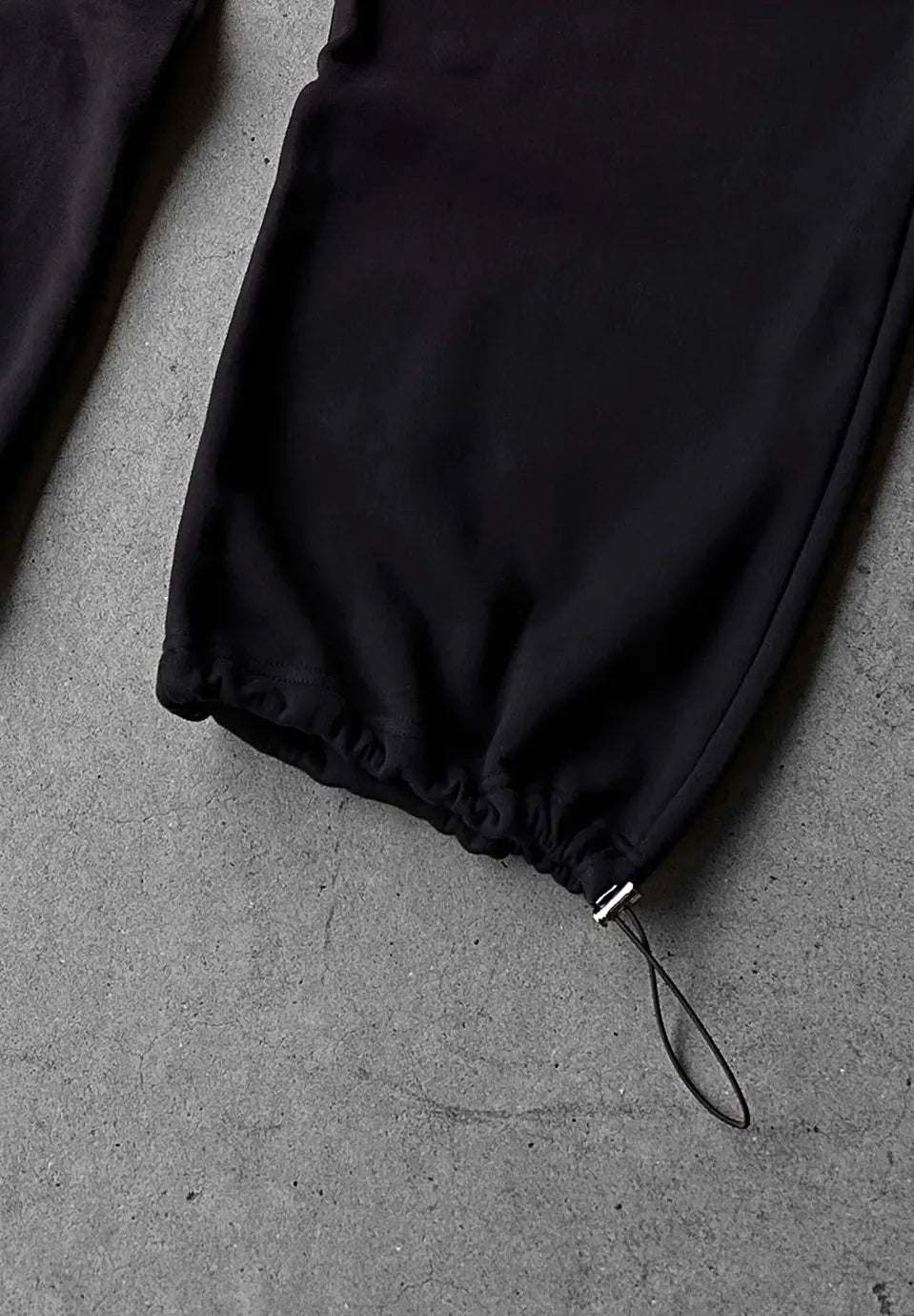 Close-up of black drawstring pants laid flat on a grey concrete surface, showing the gathered elastic cuff and a black cord loop.