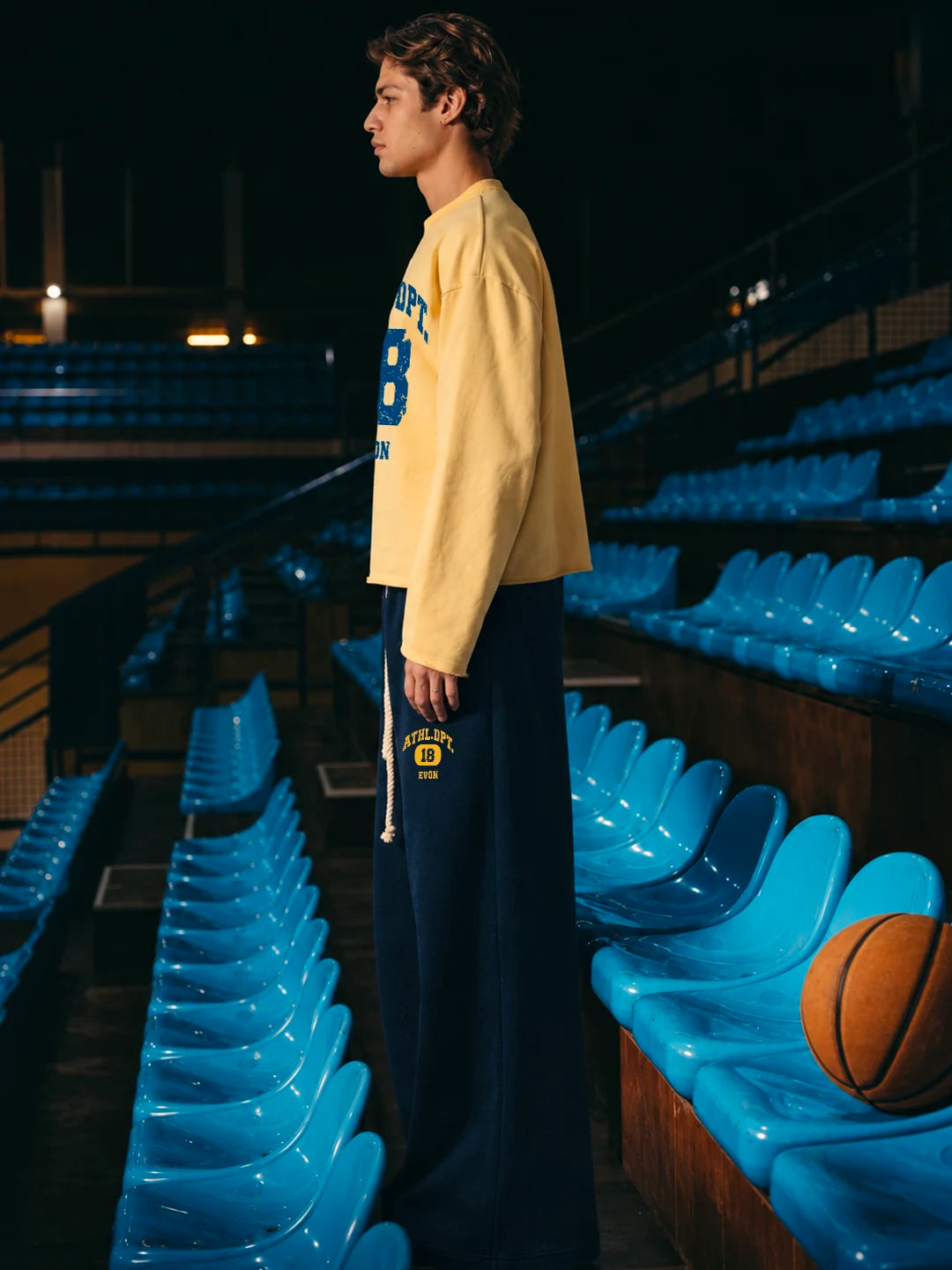 A man wearing a pale yellow long-sleeve basketball jersey with blue lettering and dark navy sweatpants. Blue stadium seats and a basketball are visible in the background.