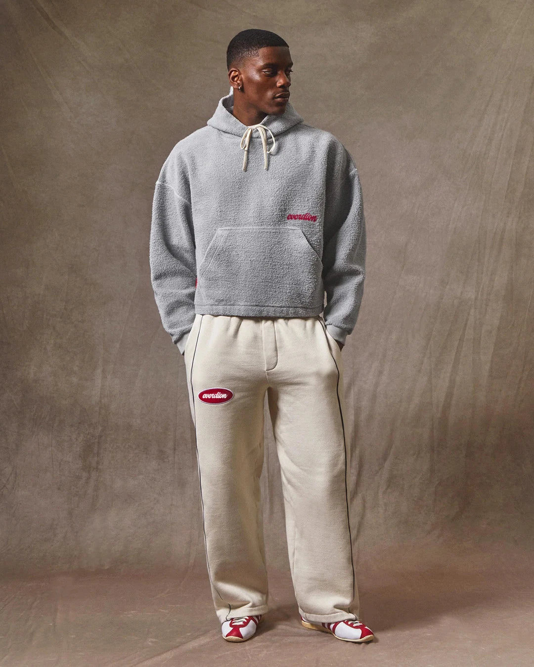 Man wearing a gray hoodie and beige pants with a brand logo against a brown background