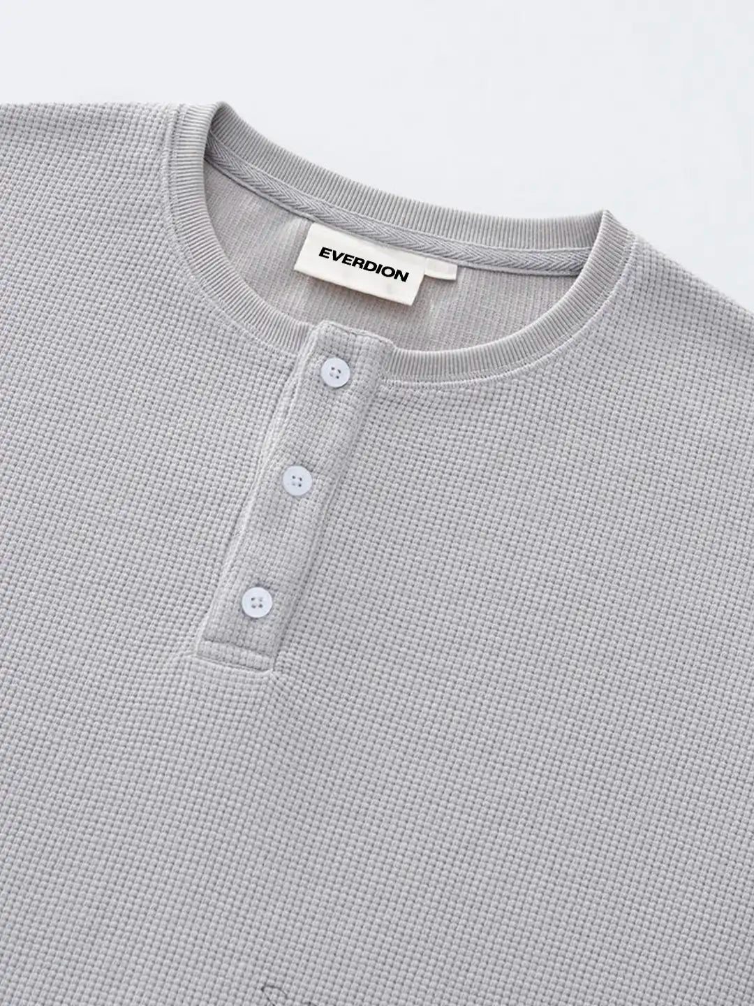 A close-up of a grey waffle-knit henley t-shirt, featuring a three-button placket and a black brand label on the inner collar.