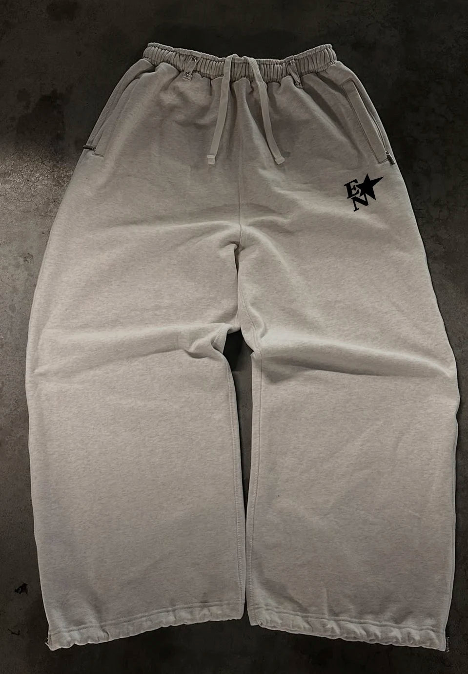 Light grey heather sweatpants with an elastic drawstring waist and a small black embroidered logo on the upper left leg, laid flat on a dark textured surface.