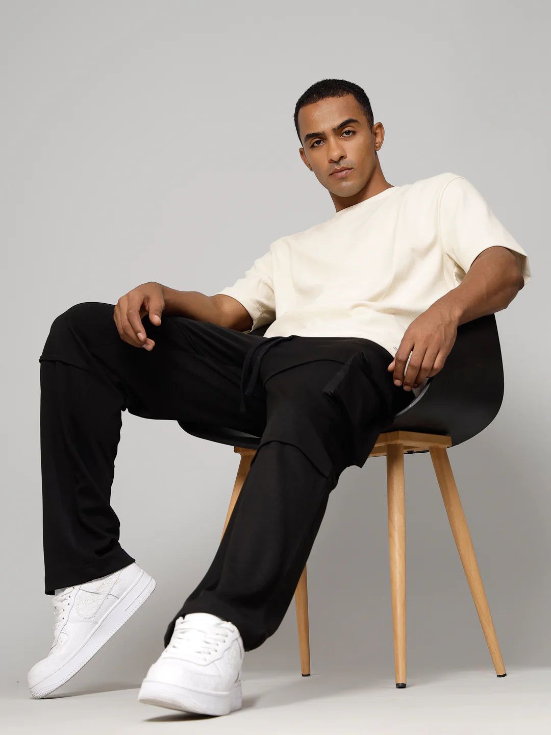 Man sitting on a chair wearing a white shirt, black pants, and white sneakers against a grey background