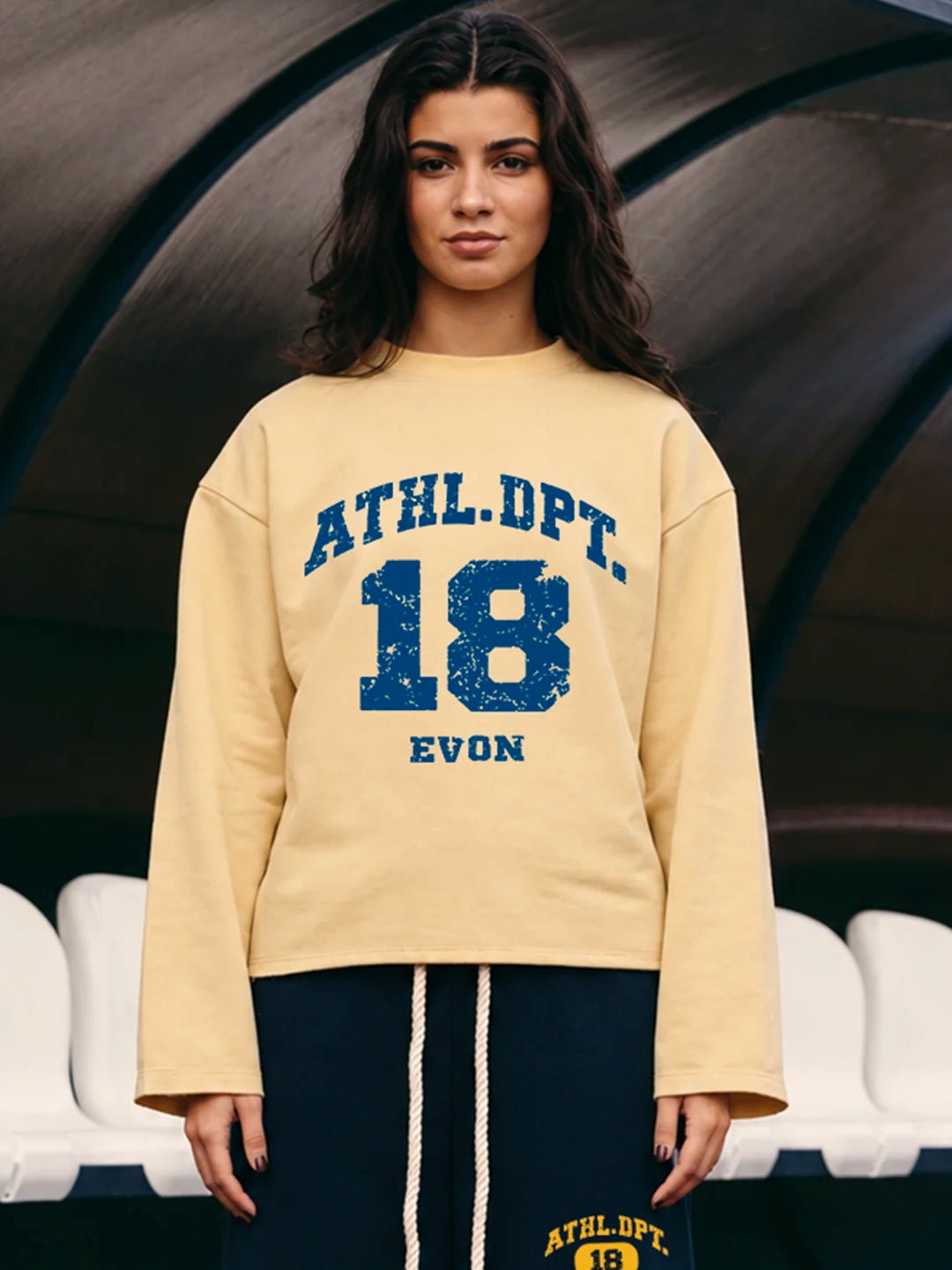 A woman wearing a cream-colored cropped sweatshirt with blue varsity-style lettering that reads 'ATHL.DPT.