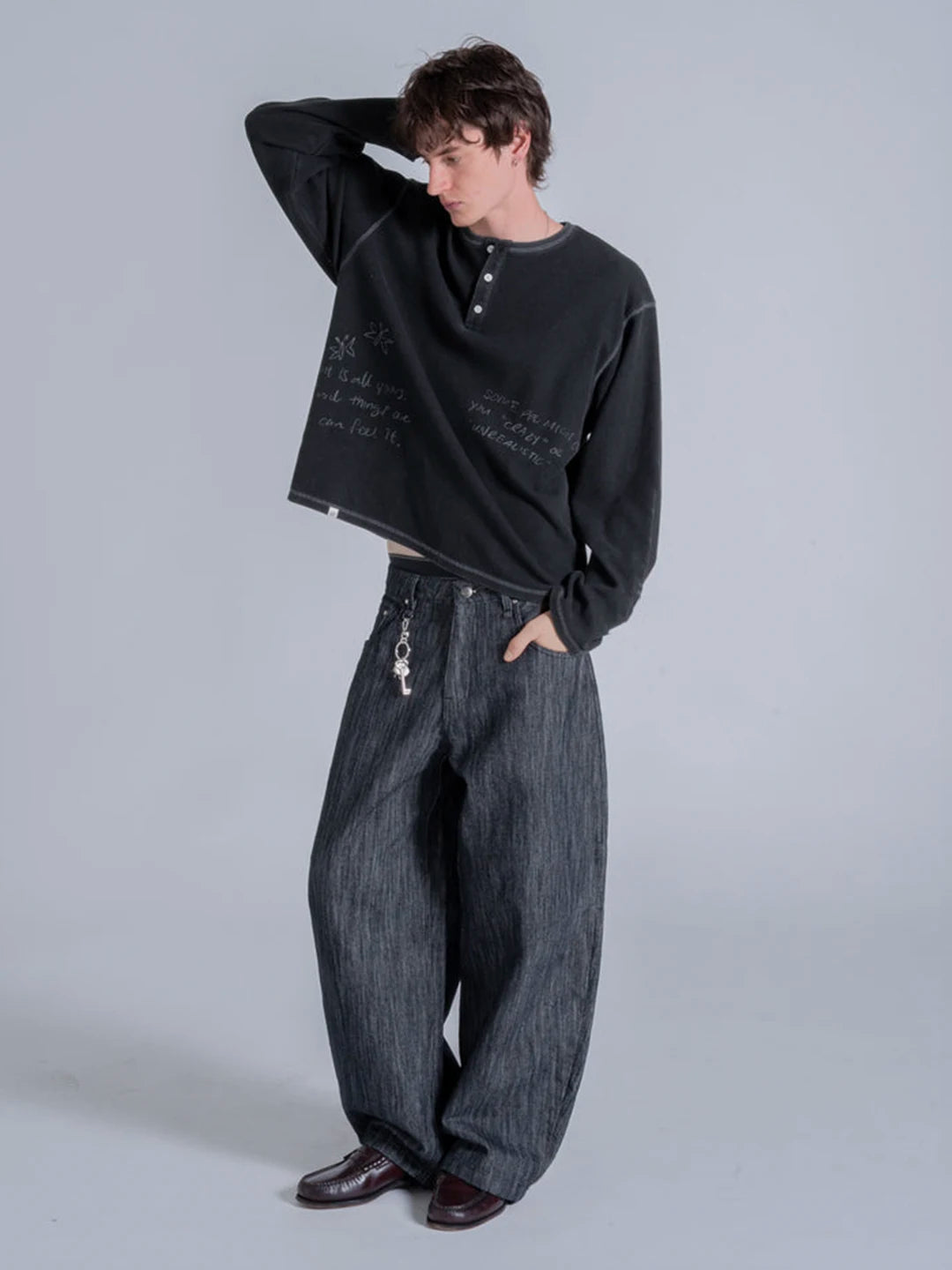 A model wearing a black long-sleeve henley tshirt with white script text, paired with wide-leg dark grey denim jeans and dark brown leather shoes