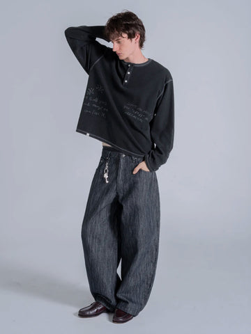 A model wearing a black long-sleeve henley tshirt with white script text, paired with wide-leg dark grey denim jeans and dark brown leather shoes