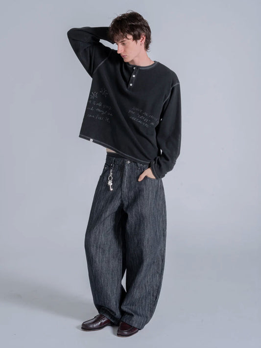 A model wearing a black long-sleeve henley tshirt with white script text, paired with wide-leg dark grey denim jeans and dark brown leather shoes