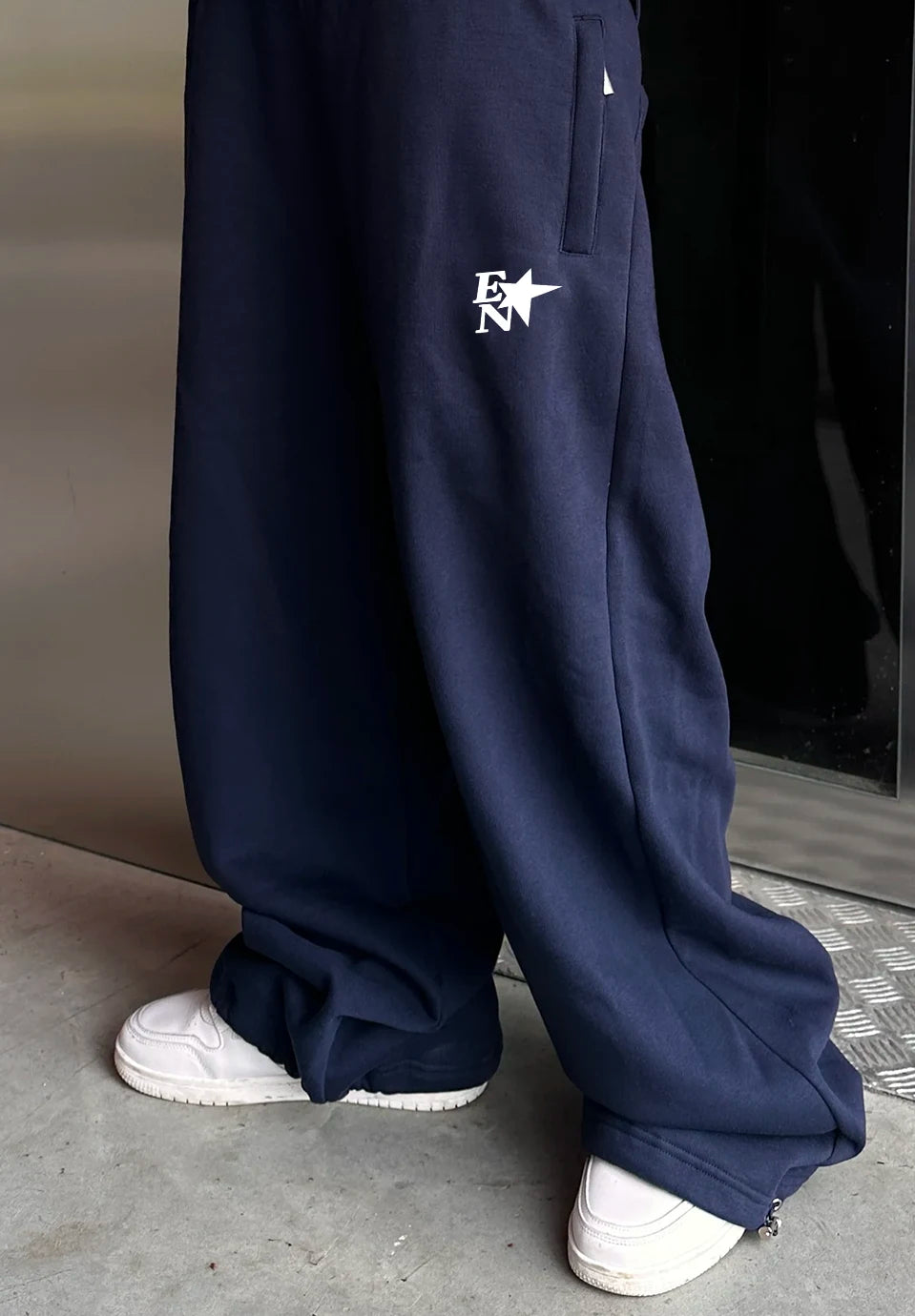 A person wearing loose navy sweatpants with a white embroidered 'EN' logo on the upper left thigh, paired with white sneakers, standing indoors near a reflective metallic surface.