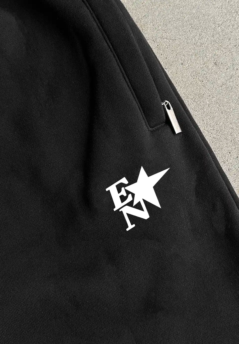 Close-up of a black fabric garment with a white stylized 'EN' logo and star on the chest, plus a small silver zipper pull