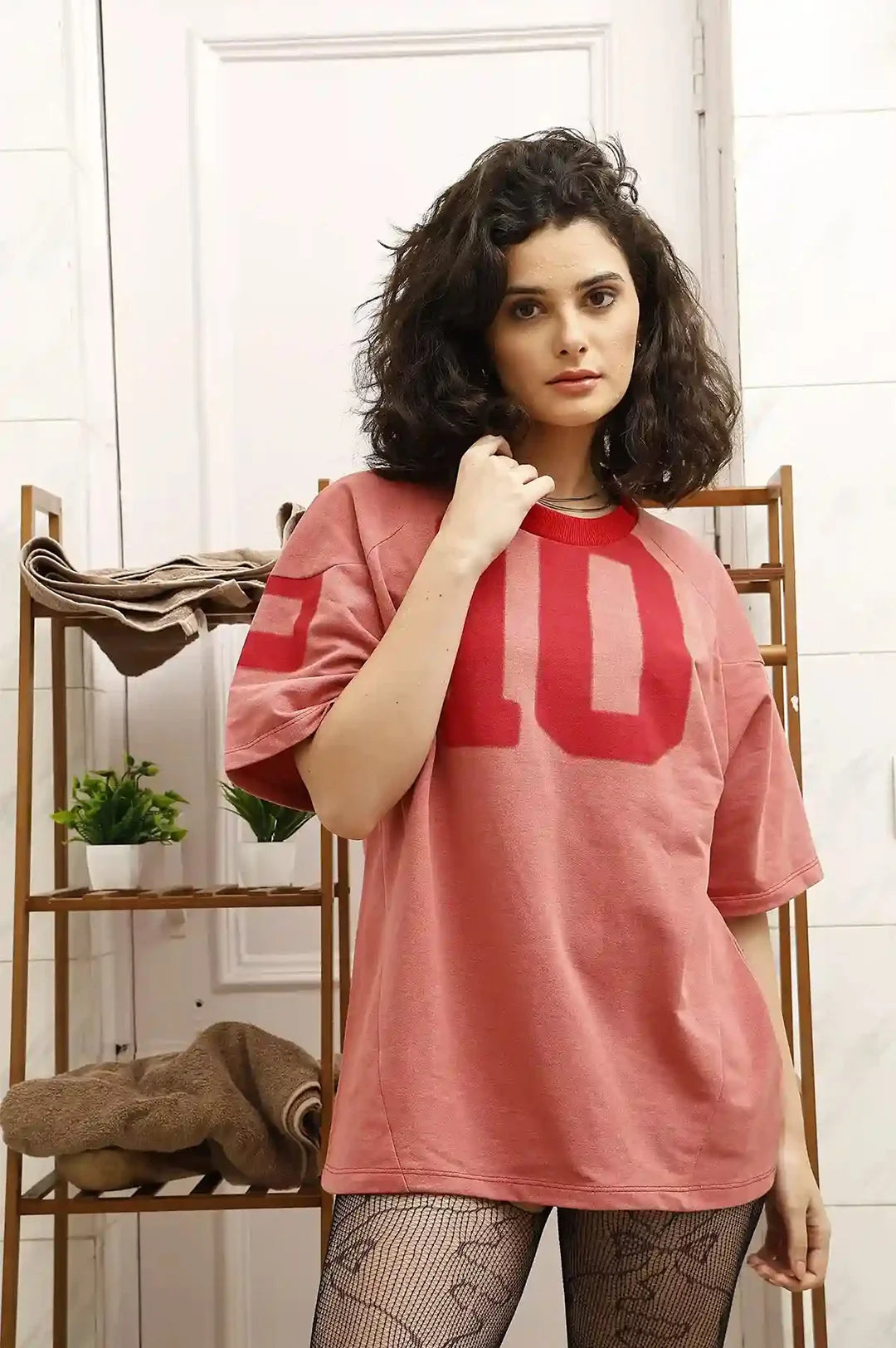 Woman wearing a pink oversized t-shirt with large red letters, standing indoors.
