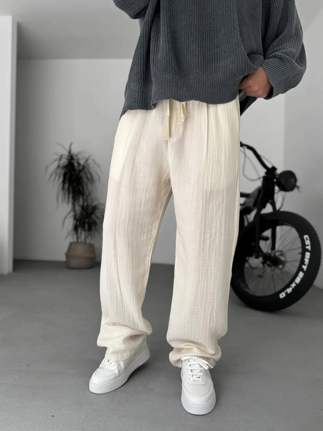 Person wearing a dark sweater and light-colored pants standing in a minimalistic room with a bicycle in the background.