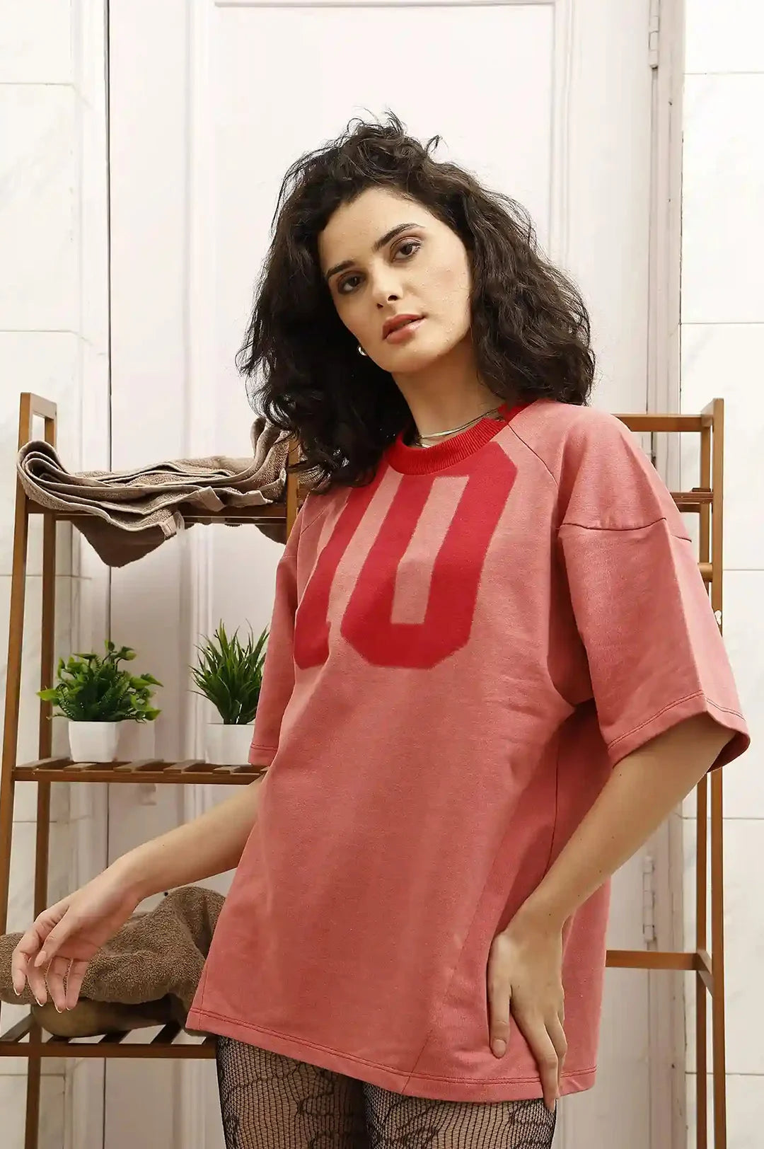 Woman wearing a pink t-shirt with a red number '10' design, standing in a room with plants and shelves.