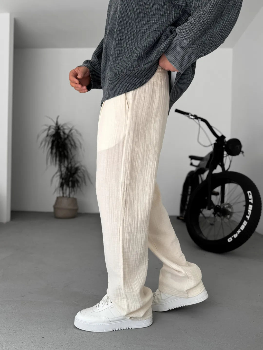 Person wearing white pants and a grey sweater in a minimalistic room with a bicycle in the background.
