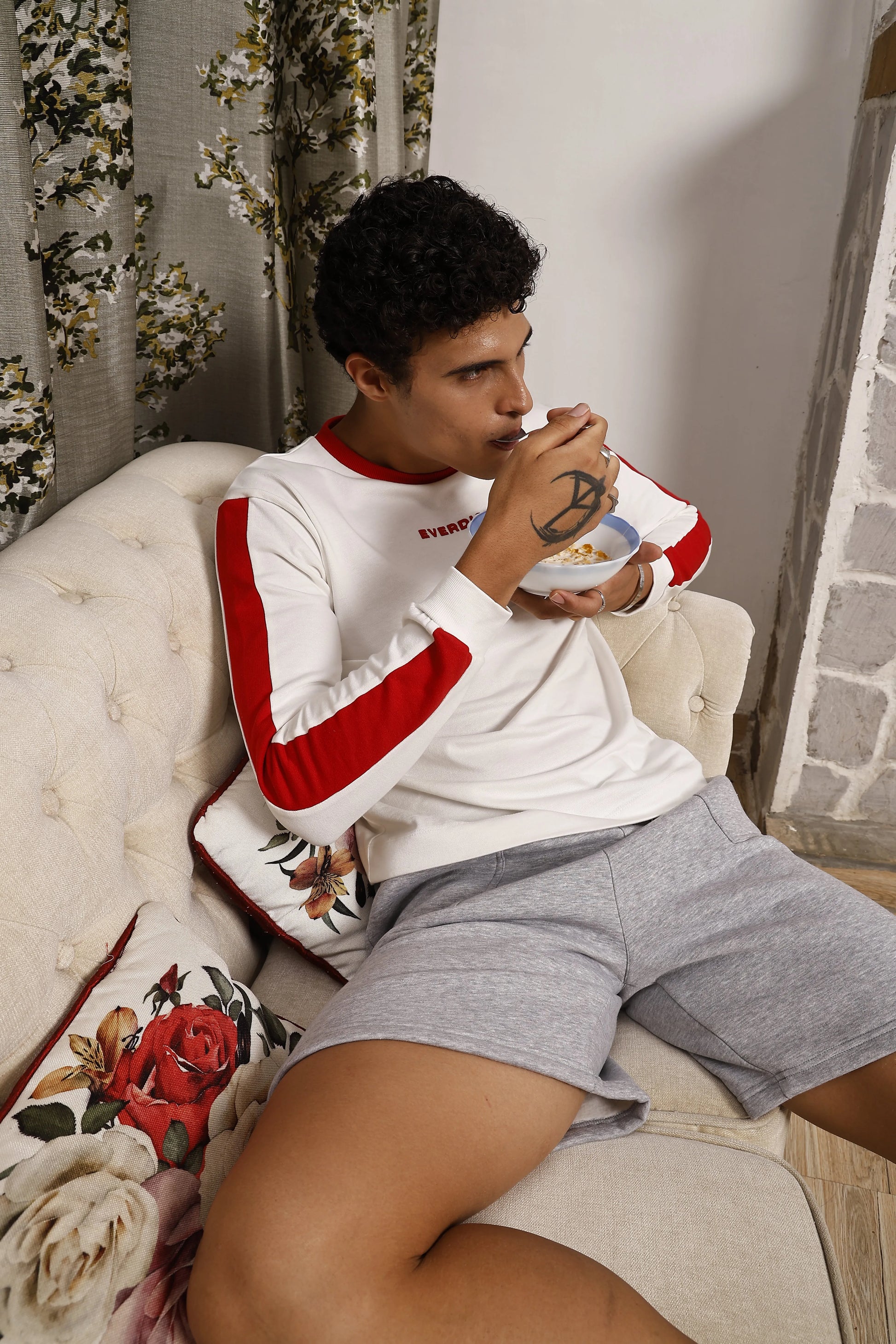 Man sitting on a couch wearing a white and red long sleeve sports t-shirt and grey shorts, holding a coffee cup.