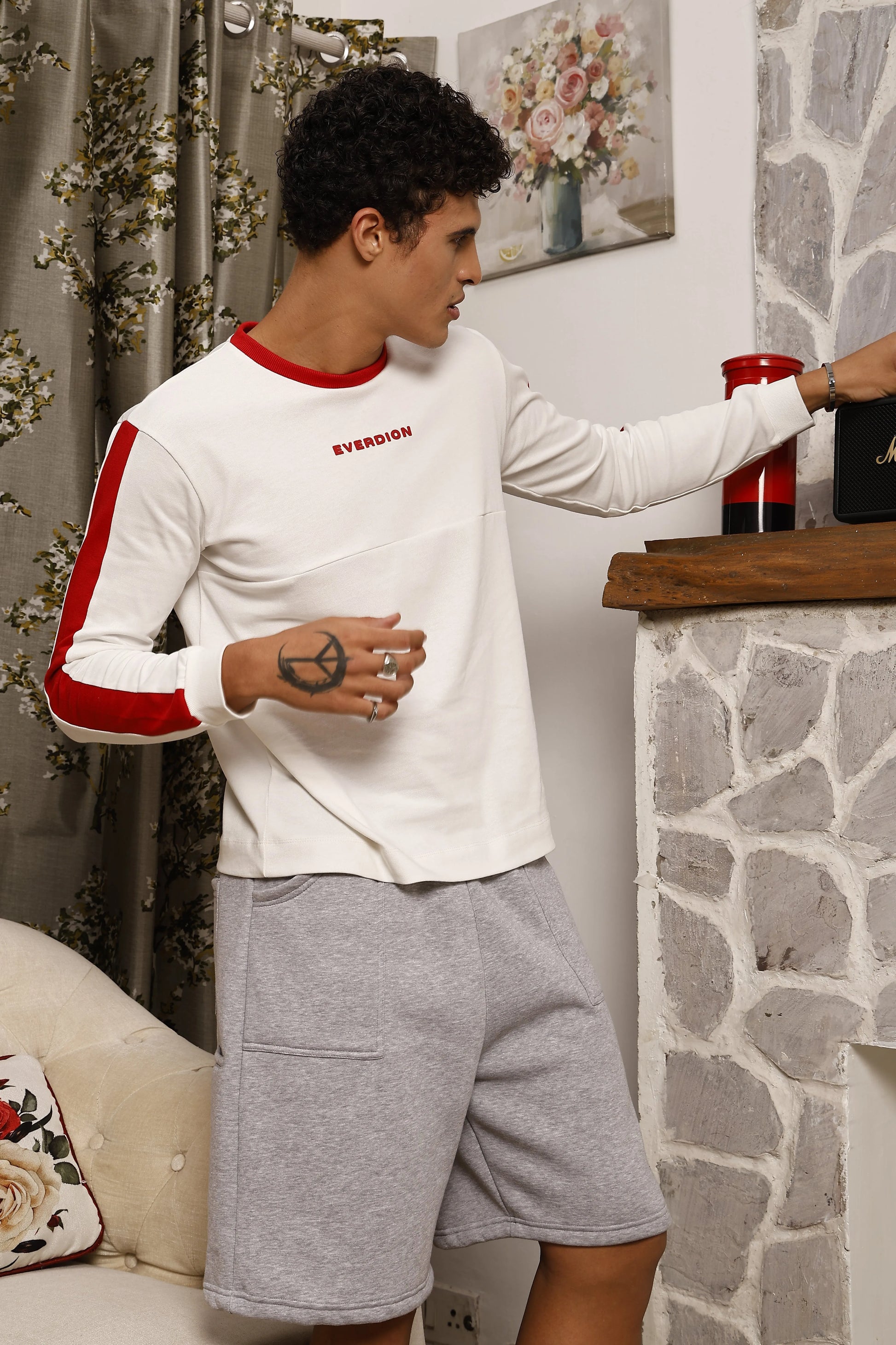 Man in a white and red long-sleeve shirt and grey shorts standing in a room with stone wall and floral decor.