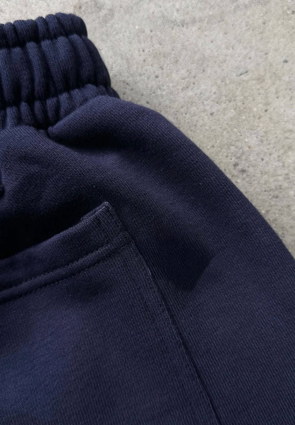 Close-up detail of dark navy blue sweatpants showing the elastic waistband and pocket seam on a grey concrete surface.