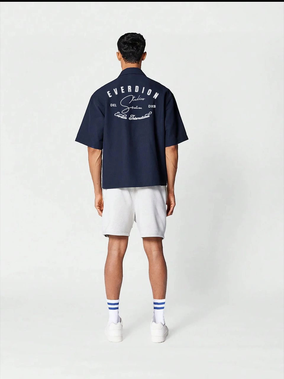 Person wearing a navy blue shirt with branding on the back, white shorts, and white shoes with blue socks.