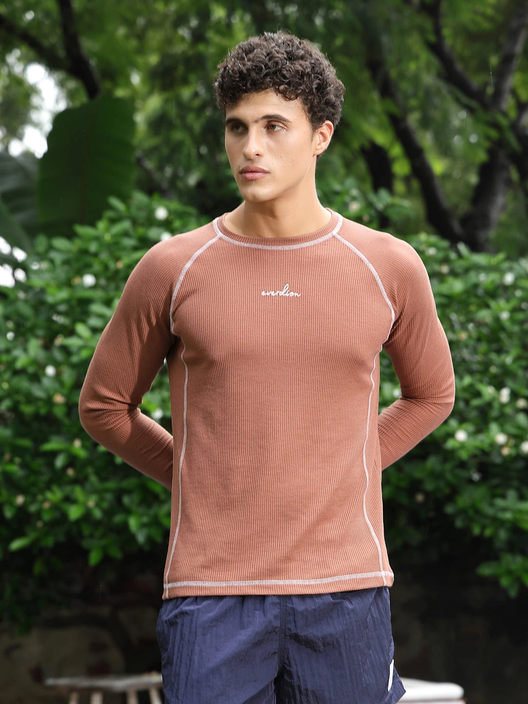 Man wearing a brown long-sleeve shirt with a brand logo, standing outdoors with greenery in the background.