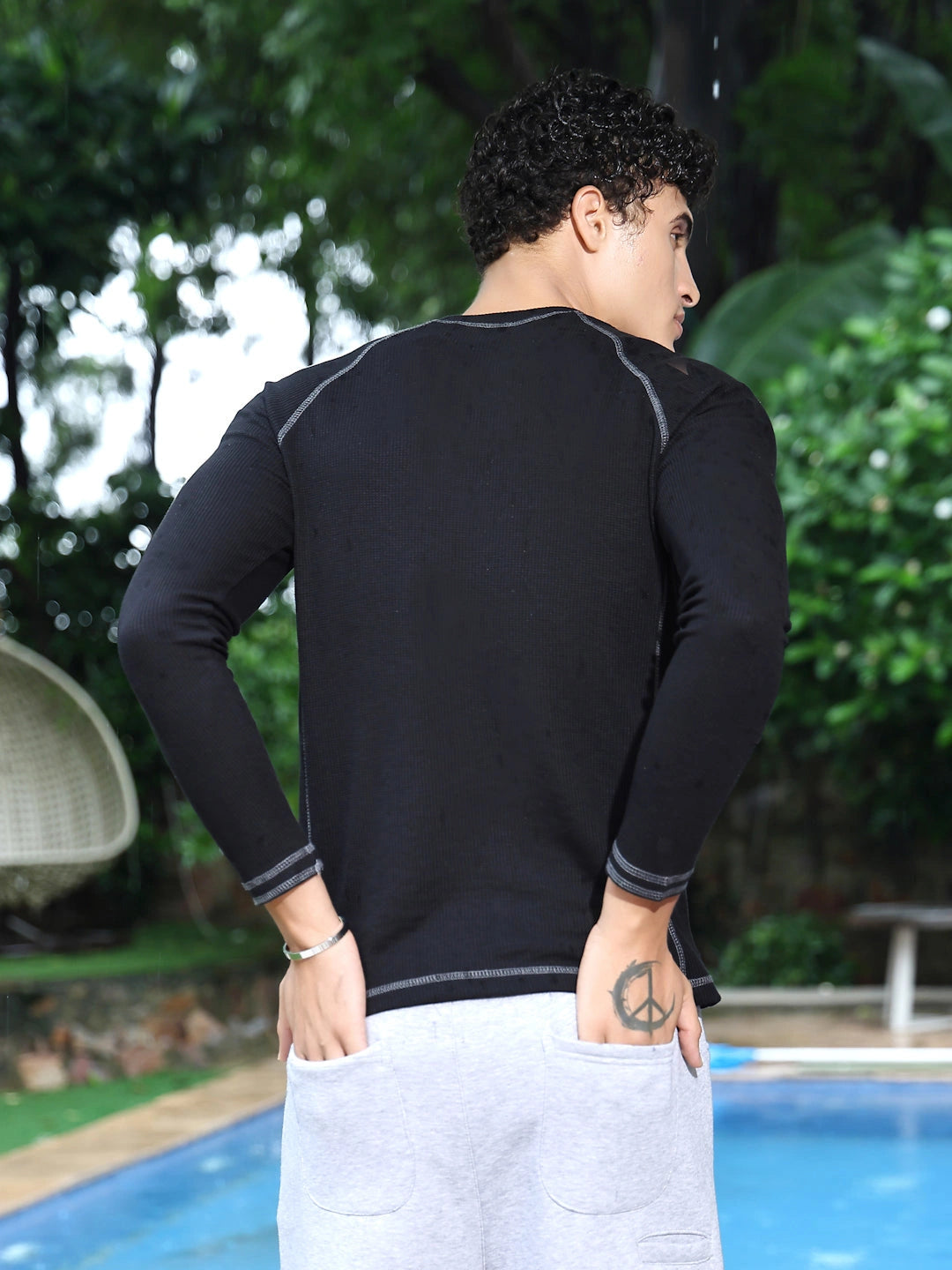 Person wearing a black long-sleeve shirt and white pants standing by a pool with greenery in the background