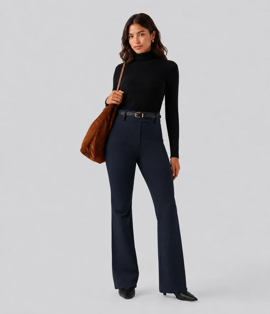 Woman wearing a black turtleneck and navy flared pants holding a brown leather handbag on a light grey background
