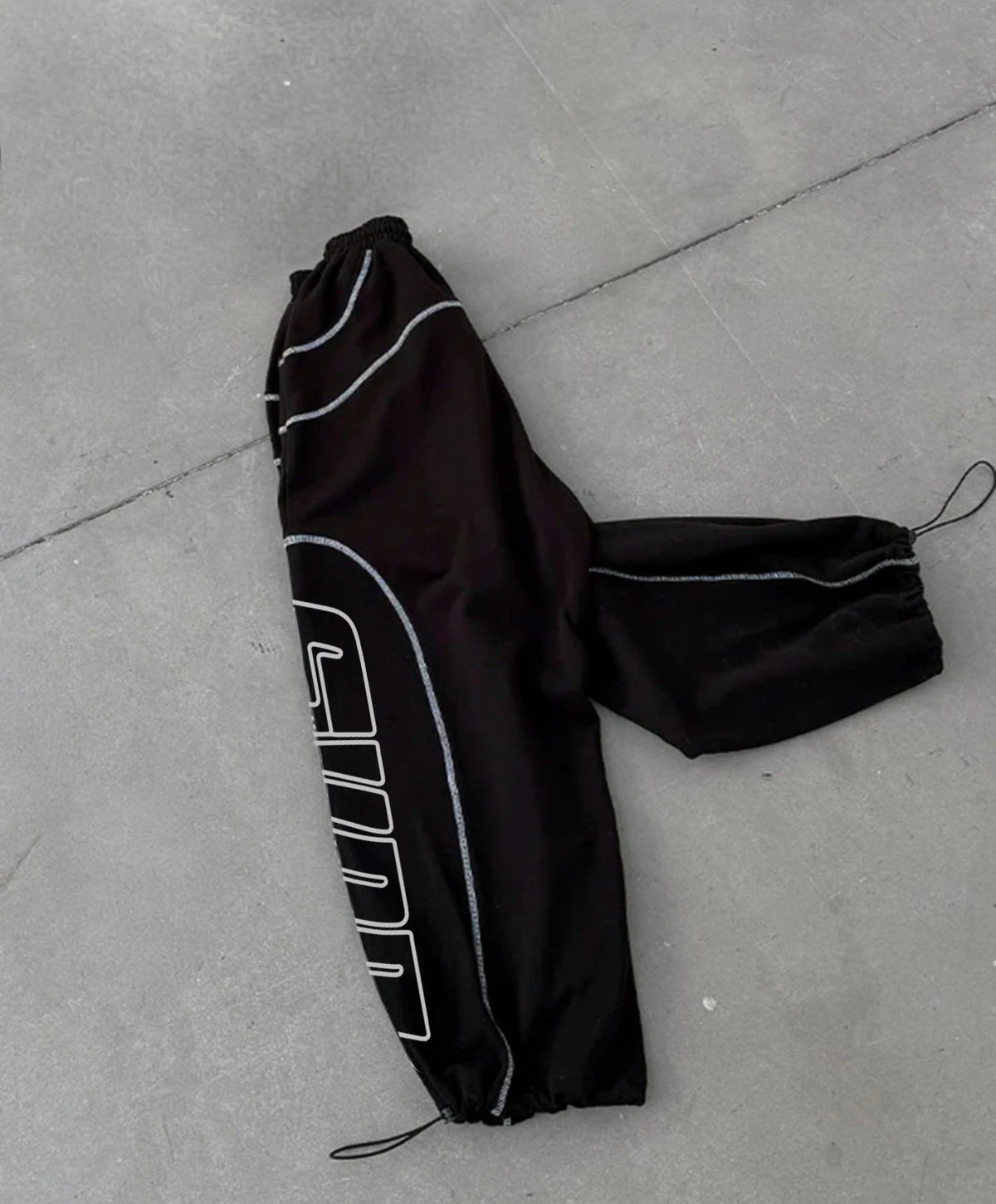 Black sweatpants with white piping and a large white outlined graphic on one leg, laid flat on a grey concrete surface.