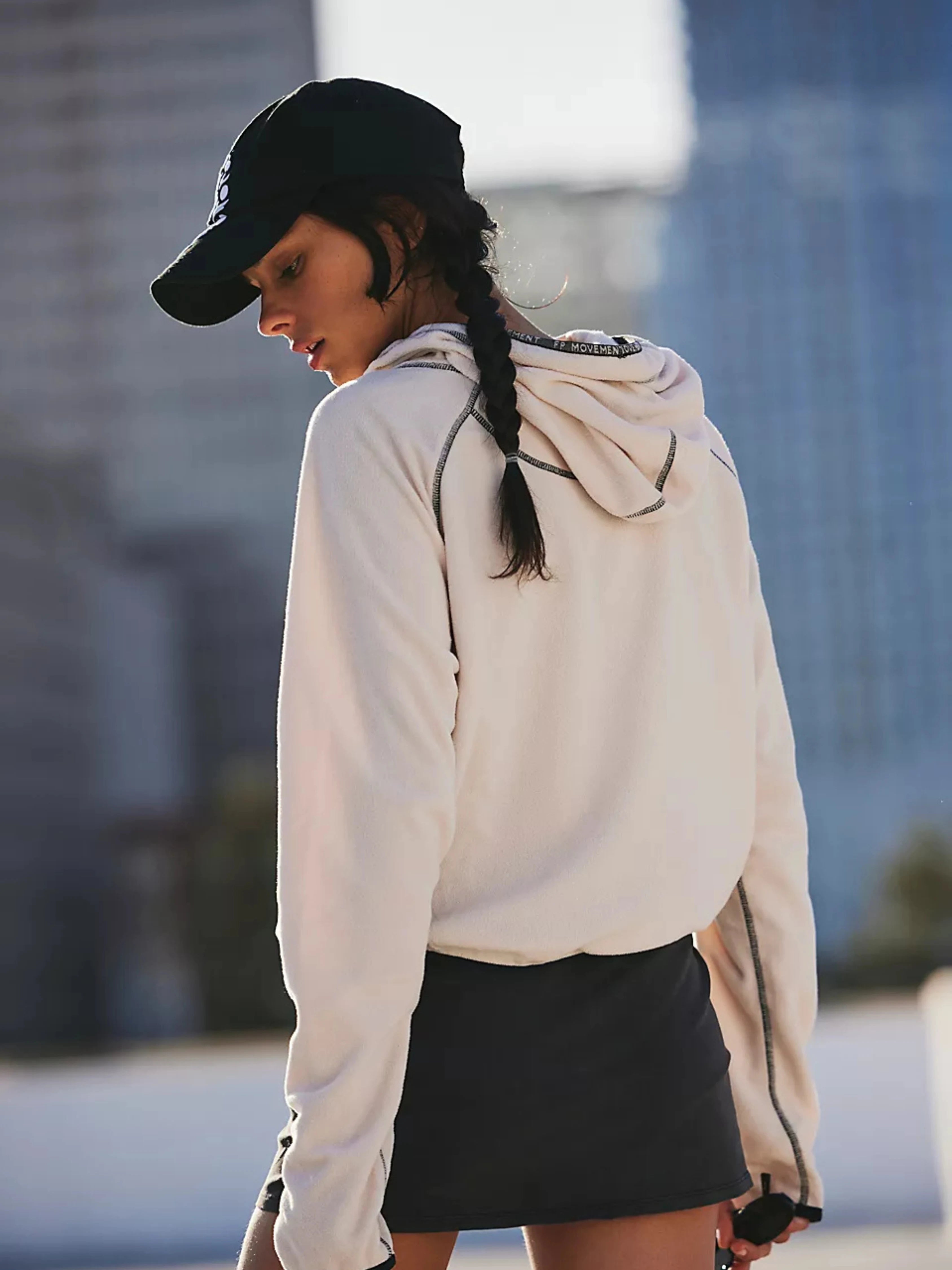 Person wearing a beige hoodie and black cap with a blurred cityscape background