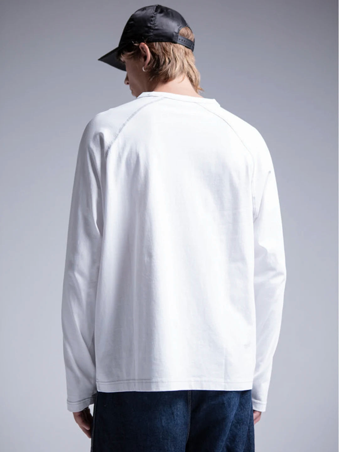A person wearing a plain white long-sleeve raglan T-shirt, dark blue jeans, and a black baseball cap