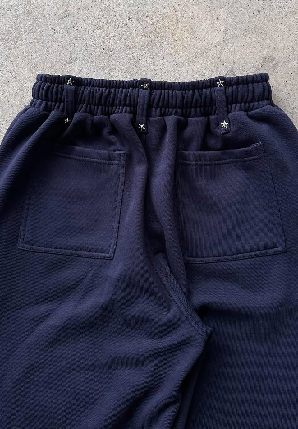 Close-up of the back waistband and pockets of dark navy sweatpants, showing an elastic gathered waist with small metallic star details and two large patch pockets.