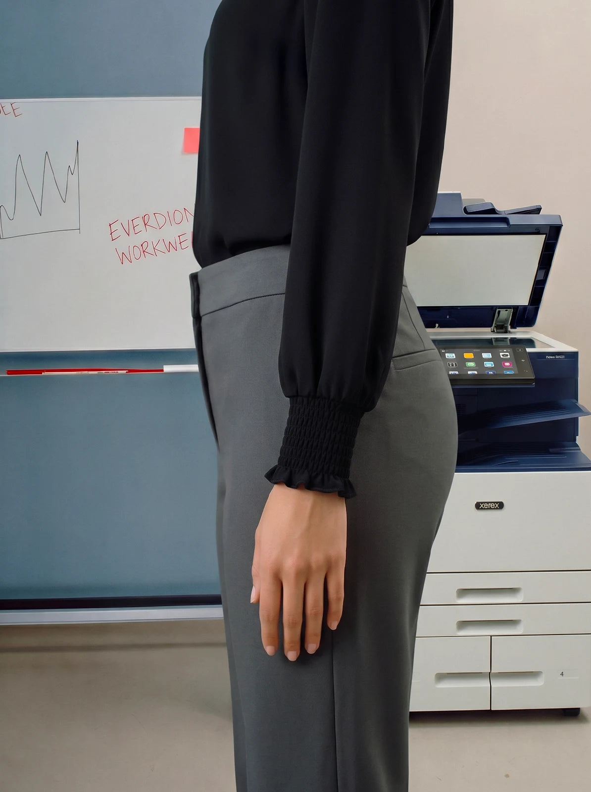 Person wearing a black shirt and grey pants in an office setting with a printer and whiteboard in the background.