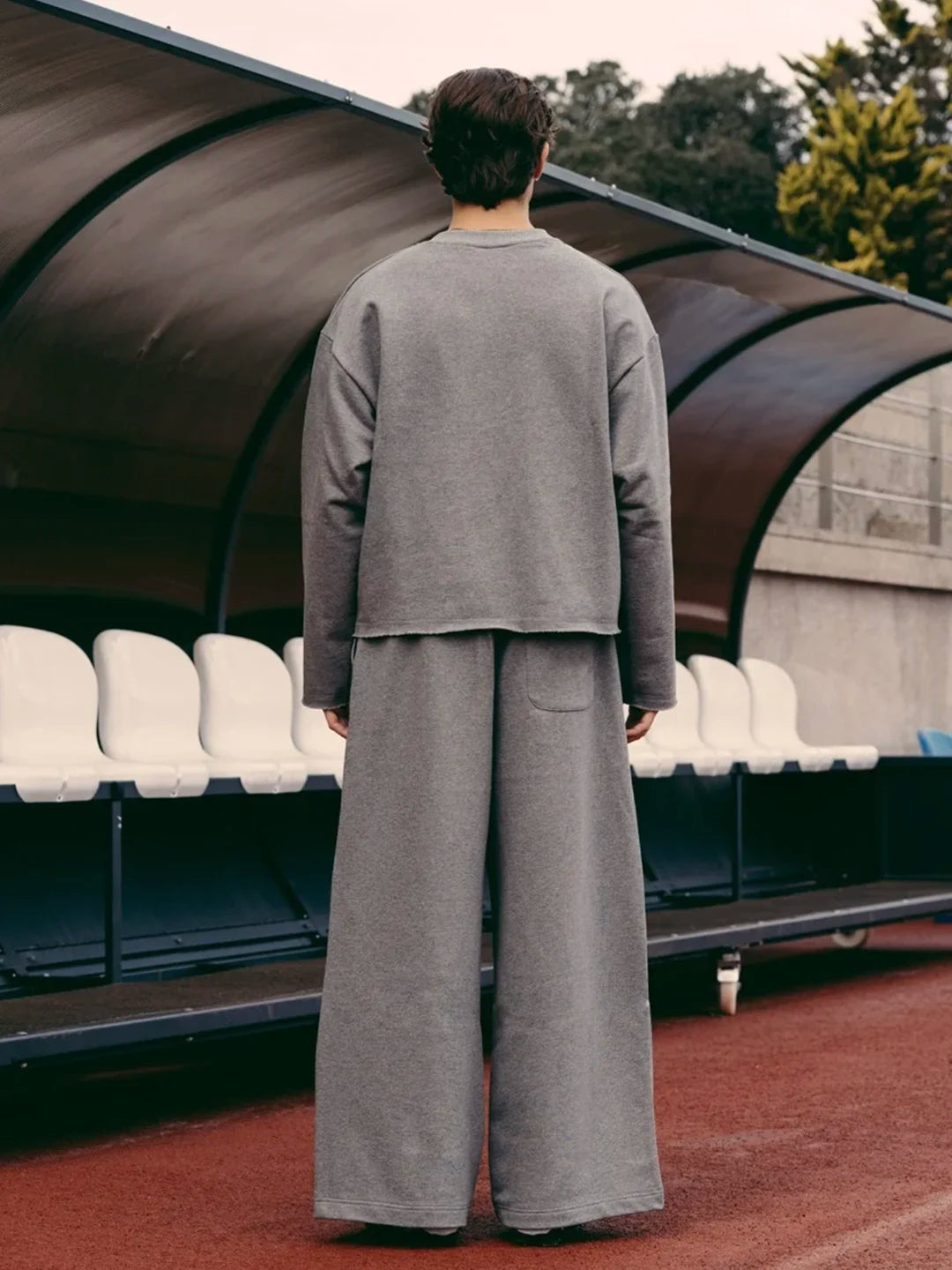 A person wearing a matching grey cropped long-sleeve top and wide-leg trousers, standing in front of stadium seating under a covered structure.