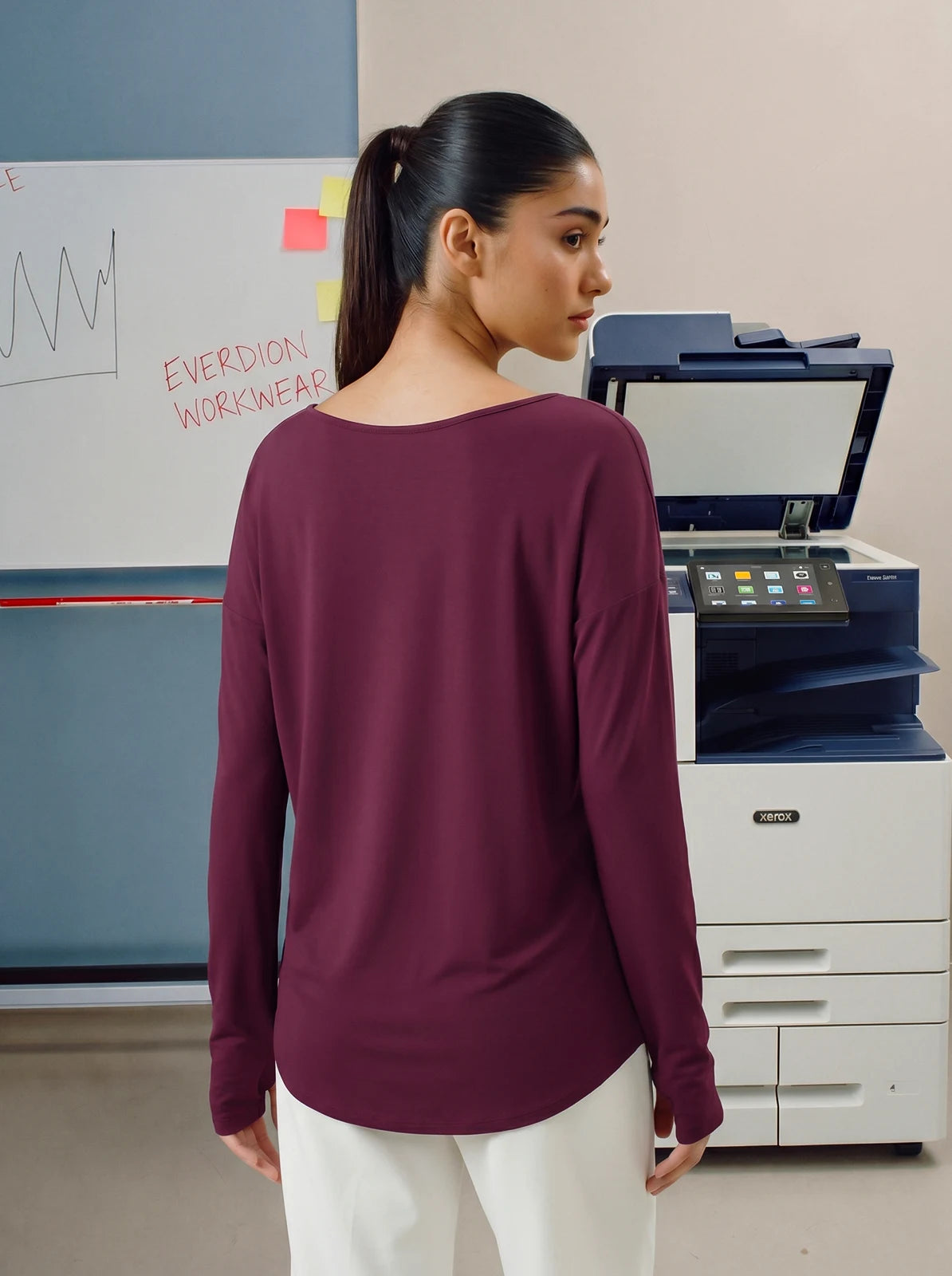 Woman in a maroon shirt standing in front of a printer and whiteboard.