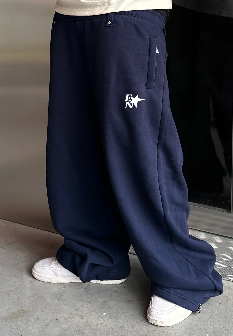 A person wearing loose navy sweatpants with a white embroidered logo on the left thigh, paired with white sneakers, shown from the waist down in an indoor setting.