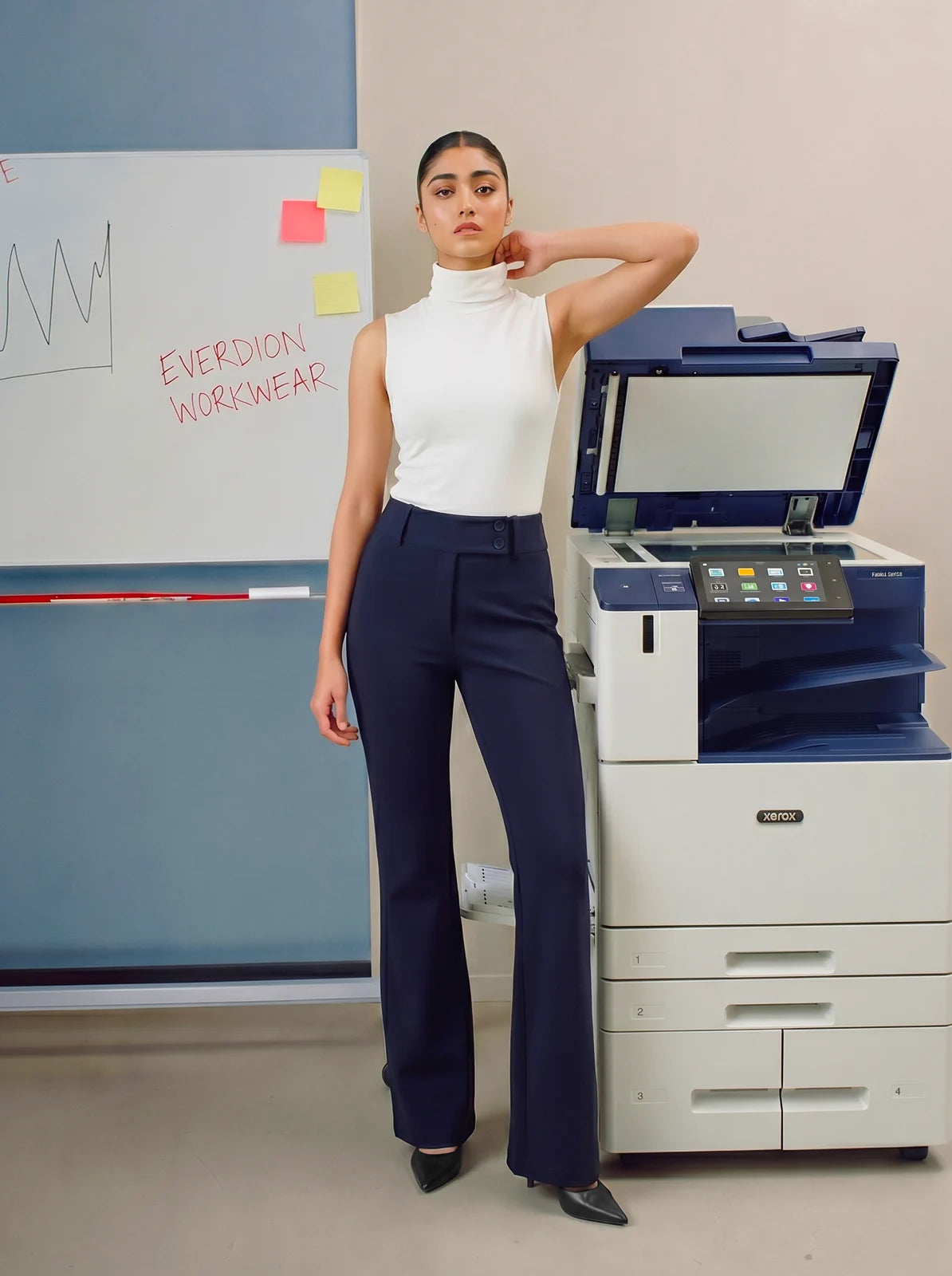 Woman in a white top and navy pants standing next to a printer with a whiteboard in the background.