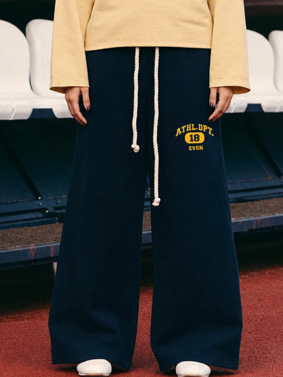 A person wearing navy wide-leg sweatpants with a cream drawstring and yellow 'ATHLET. EVON' text on the left leg, paired with a pale yellow top and white shoes, standing on a red surface in front of stadium seating.