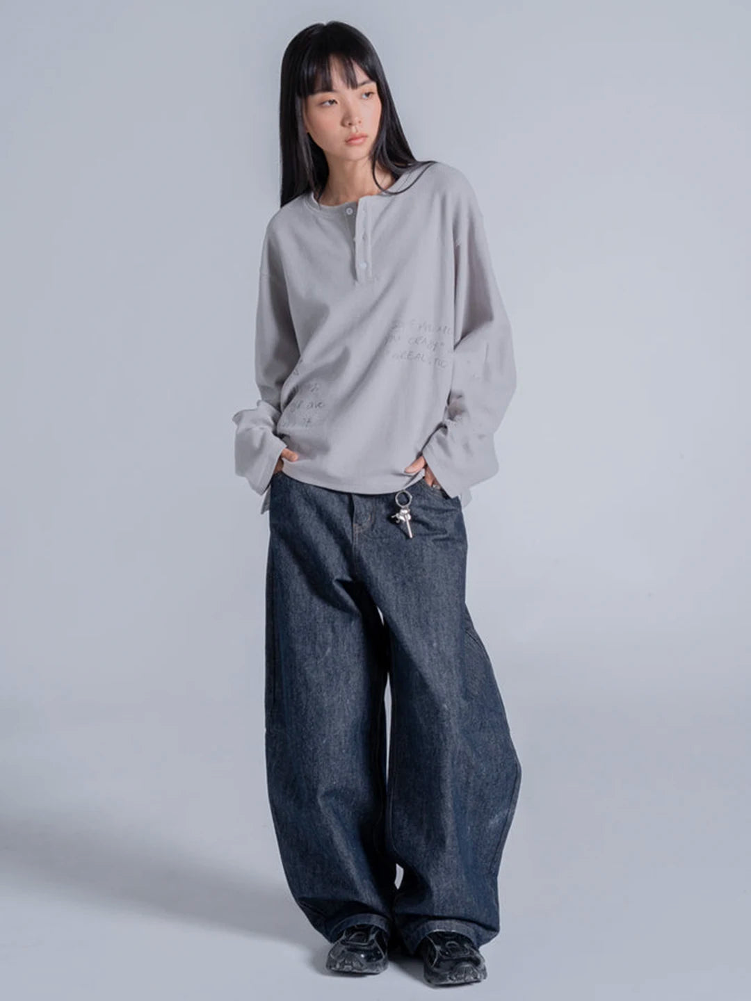 A model wearing a light grey long-sleeve henley-style tshirt with dark wide-leg denim jeans and black shoes.